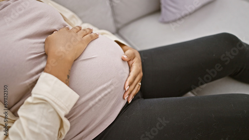 Pregnant young woman, carefree and friendly, touching her belly and massaging while sitting on the sofa in her cozy living room, immersed in the joyous emotion of expecting motherhood