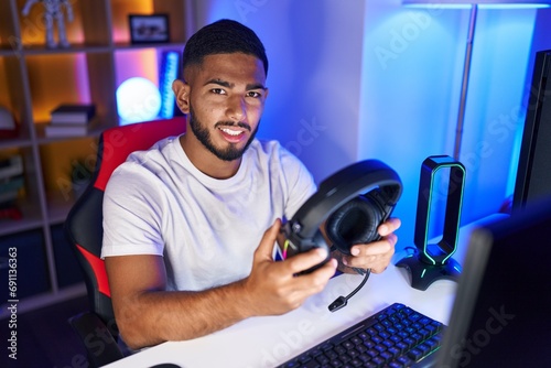 Young latin man streamer smiling confident holding headphones at gaming room