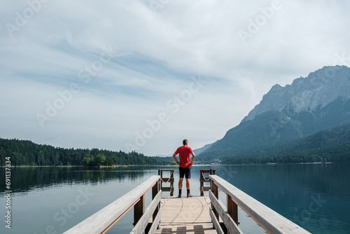 View from the shore of Lake Eibsee in Germany, with a hiker in a red shirt standing on a footbridge and looking into the distance.