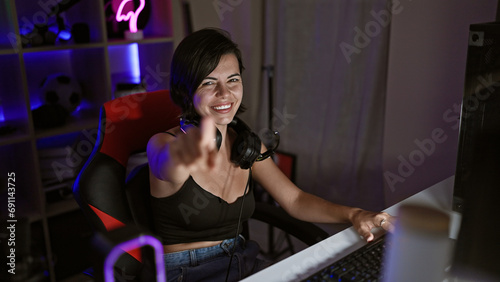 Captivating hispanic gamer gal beams, yanking off her headphones in her home gaming room, playfully pointing at you through the live stream camera photo