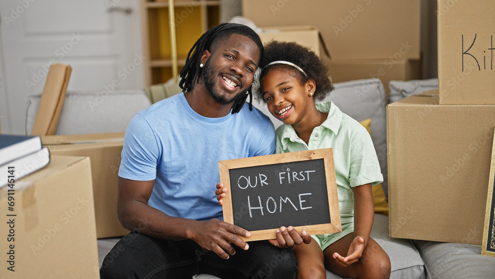 African american father and daughter holding blackboard sitting on sofa at new home - obrazy, fototapety, plakaty 
