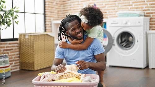 African american father and daughter hugging each other holding clothes of basket sitting on floor at laundry room photo