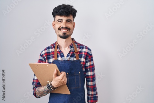 Young hispanic man with beard wearing waiter apron holding clipboard with a happy and cool smile on face. lucky person.