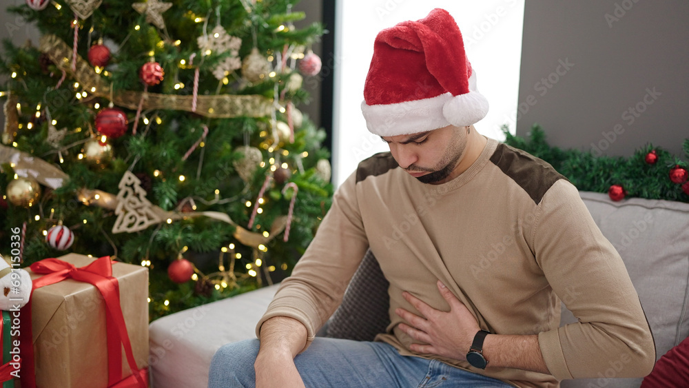 Young arab man suffering stomach ache sitting by christmas tree at home Young arab man suffering stomach ache sitting by christmas tree at home