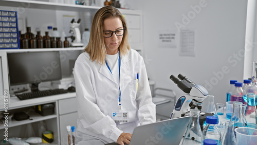 Dedicated young blonde scientist concentrates on her laptop  engrossed in vital medical research in the bustling heart of a vibrant lab