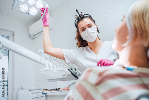 Dental clinic patient appointment in modern medical ward. Dentist doctor in magnifying glasses pointing dental light at young Female ready for teeth surgery. Health care and medicare industry image.