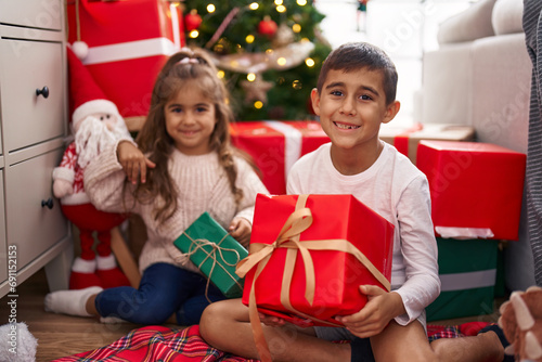 Brother and sister holding gift sitting on floor by christmas gifts at home