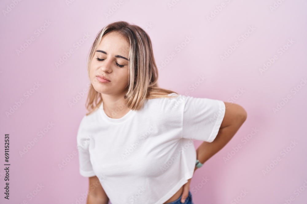 Young blonde woman standing over pink background suffering of backache, touching back with hand, muscular pain Young blonde woman standing over pink background suffering of backache, touching back with hand, muscular pain