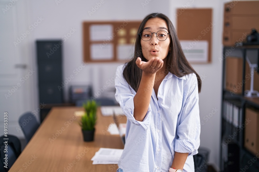 Young hispanic woman at the office looking at the camera blowing a kiss with hand on air being lovely and sexy. love expression. Young hispanic woman at the office looking at the camera blowing a kiss with hand on air being lovely and sexy. love expression.