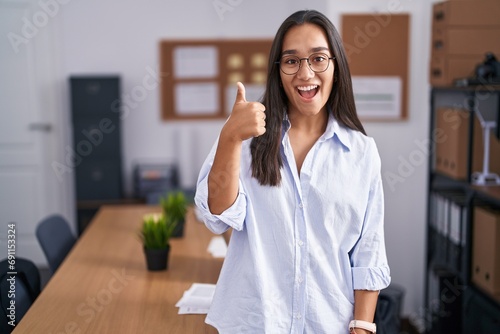 Young hispanic woman at the office doing happy thumbs up gesture with hand. approving expression looking at the camera showing success. photo
