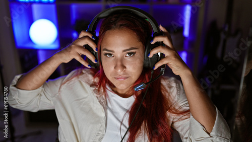 Focused young redhead woman streamer, engrossed in intense gaming session, in a serious virtual face-off, headset on, encapsulated in the dimly-lit indoors of her home gaming room