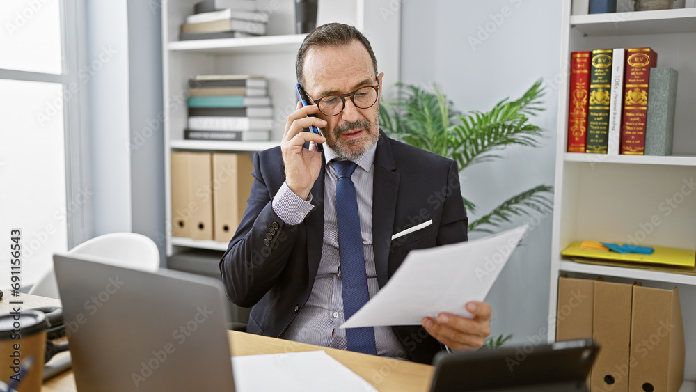 Energetic middle age man with grey hair, immersed in his professional role at the office, confidently talking on a smartphone while attentively reading a crucial business document. Energetic middle age man with grey hair, immersed in his professional role at the office, confidently talking on a smartphone while attentively reading a crucial business document.