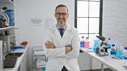 Happy mature, grey-haired male scientist with a confident smile enjoys his research work, sitting at his lab table, arms crossed in a welcoming gesture, connected with science and medicine. photo