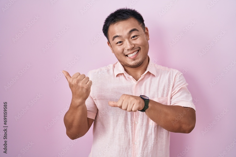 Chinese young man standing over pink background pointing to the back behind with hand and thumbs up, smiling confident Chinese young man standing over pink background pointing to the back behind with hand and thumbs up, smiling confident