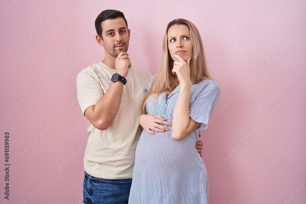 Young couple expecting a baby standing over pink background looking confident at the camera smiling with crossed arms and hand raised on chin. thinking positive. Young couple expecting a baby standing over pink background looking confident at the camera smiling with crossed arms and hand raised on chin. thinking positive.