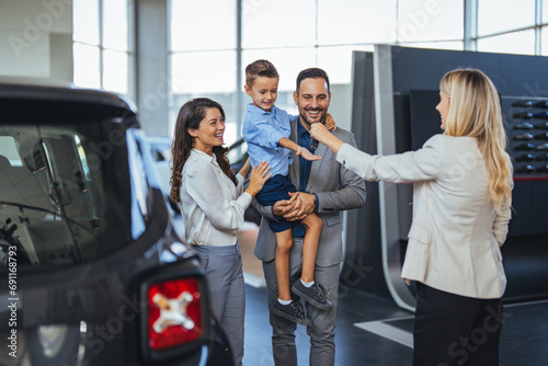 Happy family choosing new car, salesman showing them luxury auto at automobile dealership store. Customers selecting vehicle, consulting manager at modern showroom shop