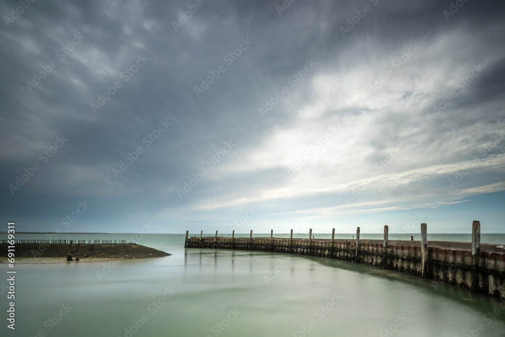 Obraz premium Urbex harbor in a round shape of weathered concrete with rusted boolders and rotten mooring posts and above a threatening cloudy sky