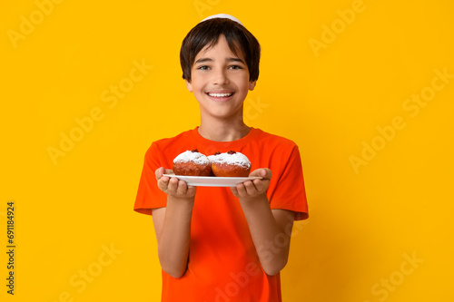 Little boy in kipa holding plate with tasty donuts on yellow background. Hanukkah celebration photo