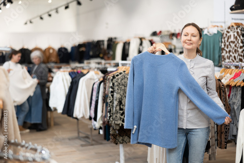 Happy middle-aged woman buyer choosing convenient jumper in clothing shop with large assortment