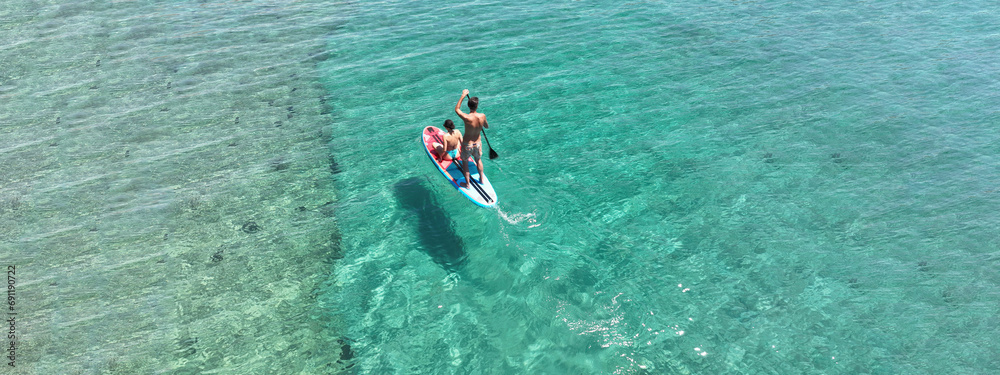 Aerial drone ultra wide photo of couple sitting on SUP or Stand Up Paddle board cruising in low speed in tropical paradise exotic bay with emerald crystal clear calm sea