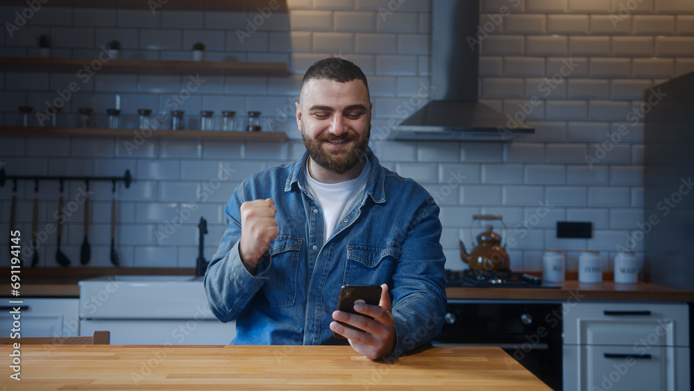 Bearded young man sitting against the kitchen counter checking messages read notice receive good news on smartphone in e-mail celebrate great opportunity Bearded young man sitting against the kitchen counter checking messages read notice receive good news on smartphone in e-mail celebrate great opportunity