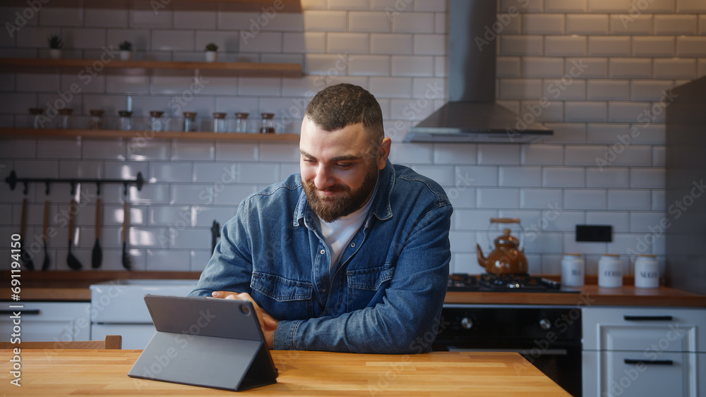 Cozy bearded young adult man sitting against the kitchen counter relaxing and using tablet device surfing internet, using social media or app Cozy bearded young adult man sitting against the kitchen counter relaxing and using tablet device surfing internet, using social media or app