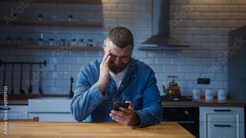 Bearded young adult man sitting against the kitchen counter having headache while using smartphone. Tired young man get having pain from a headache or migraine	 photo