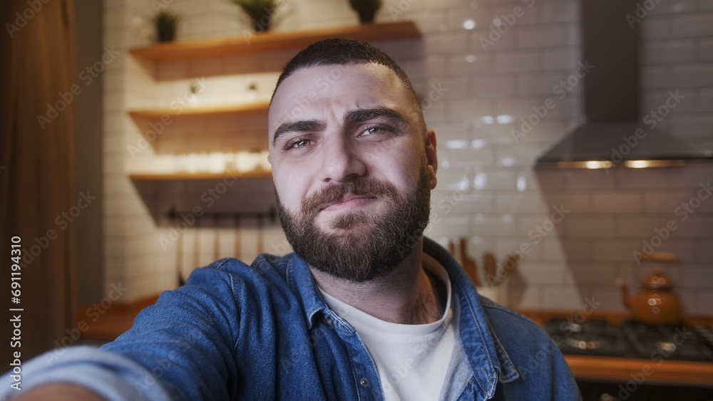 Fototapeta premium Selfie of cozy young bearded adult man holding a smartphone in hand looking at camera making video or selfie in kitchen. Modern tech, self leisure, enjoying, having fun