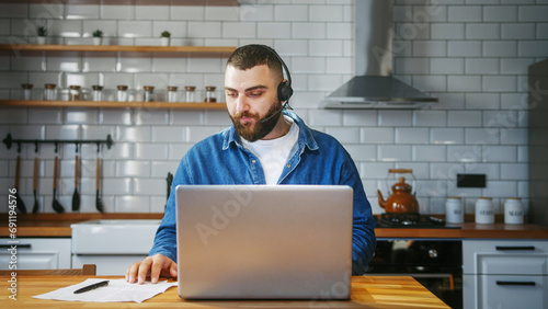 Bearded young adult business man wear wireless headset sitting against the kitchen counter having video conference calling on laptop computer talk by webcam in online chat