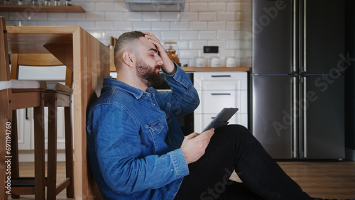 Young man sitting on the floor leaning against the kitchen using tablet device having anxiety and stress. Upset man reacting to loss, bad news