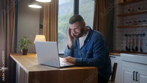 Bearded young adult man sitting against the kitchen counter having headache while using laptop. Tired young man get having pain from a headache or migraine	