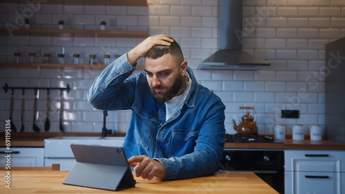Bearded young man sitting against the kitchen counter using tablet device having anxiety and stress. Upset man reacting to loss, bad news