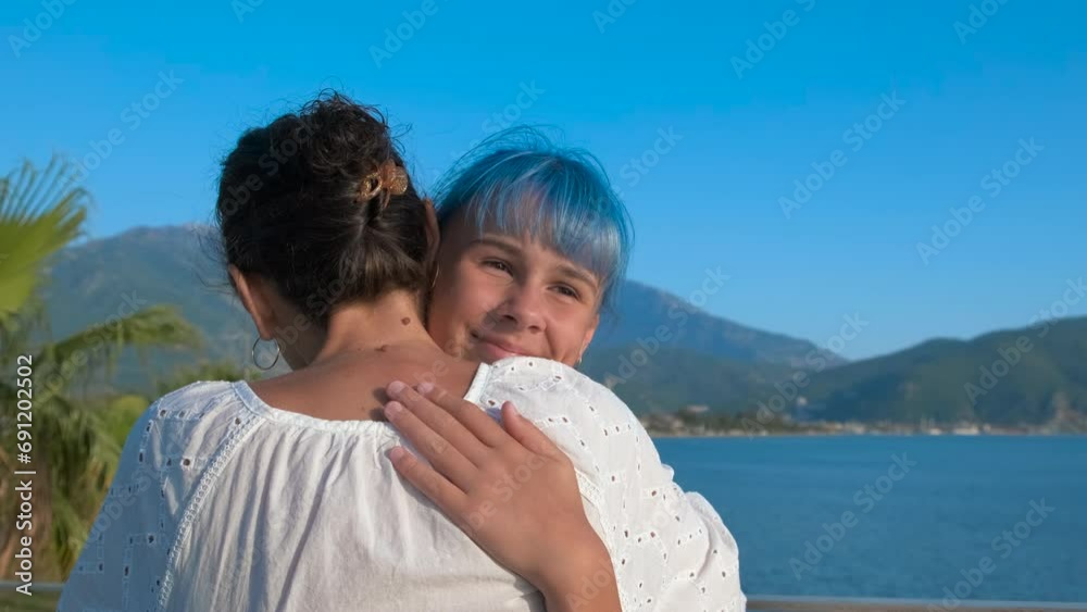 Modern teen hug mother by sea. A lovely blue haired teen girl embrace her mother on the river bank against waves in the sun light. Modern teen hug mother by sea. A lovely blue haired teen girl embrace her mother on the river bank against waves in the sun light.