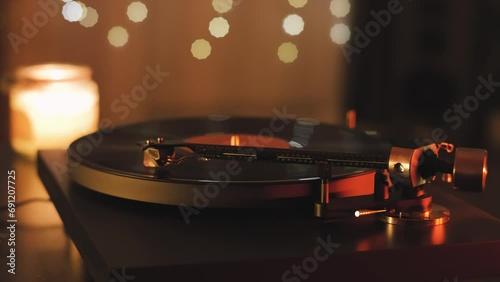 A vinyl record spins in the modern gramophone music player and plays an old disco. Close-up shot of custom vinyl turntable player with carbon tonearm. Candle and garlands in the background photo