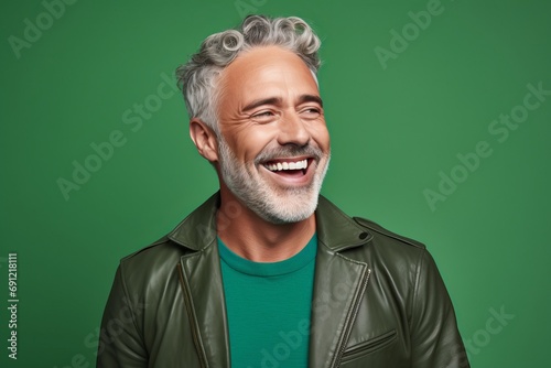 Cheerful senior man with grey hair and beard laughing while standing against green background