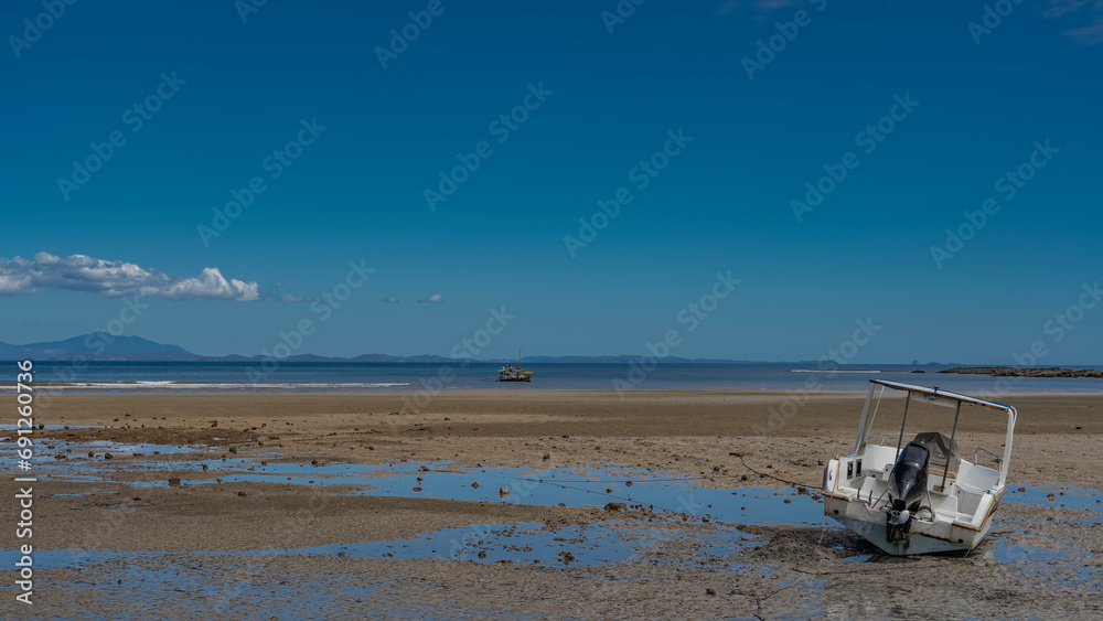 The motorboat is tilted on the bottom of the sea, exposed at low tide. Nearby there are puddles of water, scattered stones. In the distance, a sailboat with fishermen is visible in the blue ocean. 