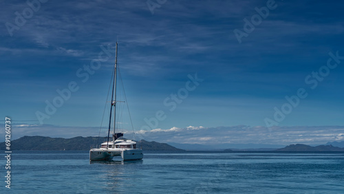 A white yacht in the blue ocean. A tall mast against the background of the sky and light clouds. The mountain range is visible in the distance. Madagascar.