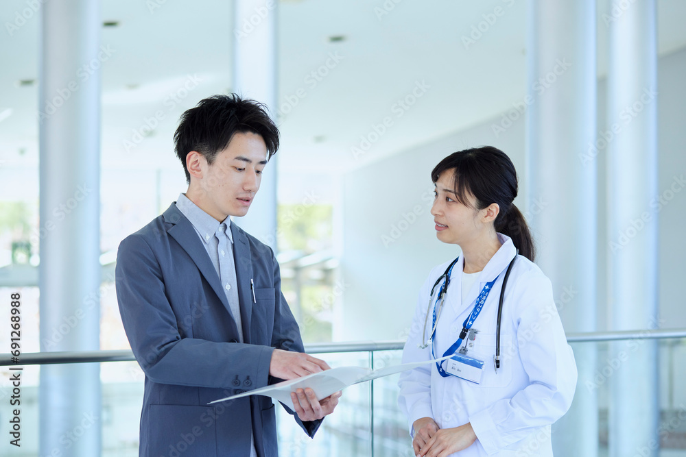 病院内にてスーツを着たMRの男性と話をする女性医師 - obrazy, fototapety, plakaty 
