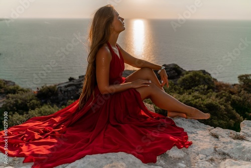 Woman sunset sea red dress, side view a happy beautiful sensual woman in a red long dress posing on a rock high above the sea on sunset.