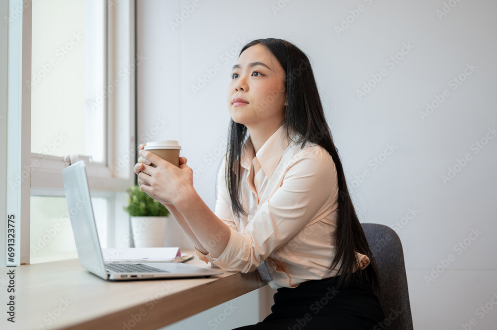 An attractive Asian businesswoman is daydreaming while enjoying her coffee in the office. An attractive Asian businesswoman is daydreaming while enjoying her coffee in the office.