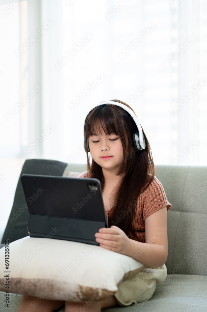 A cute young Asian girl wearing headphones, studying online on her digital tablet on a sofa. A cute young Asian girl wearing headphones, studying online on her digital tablet on a sofa.