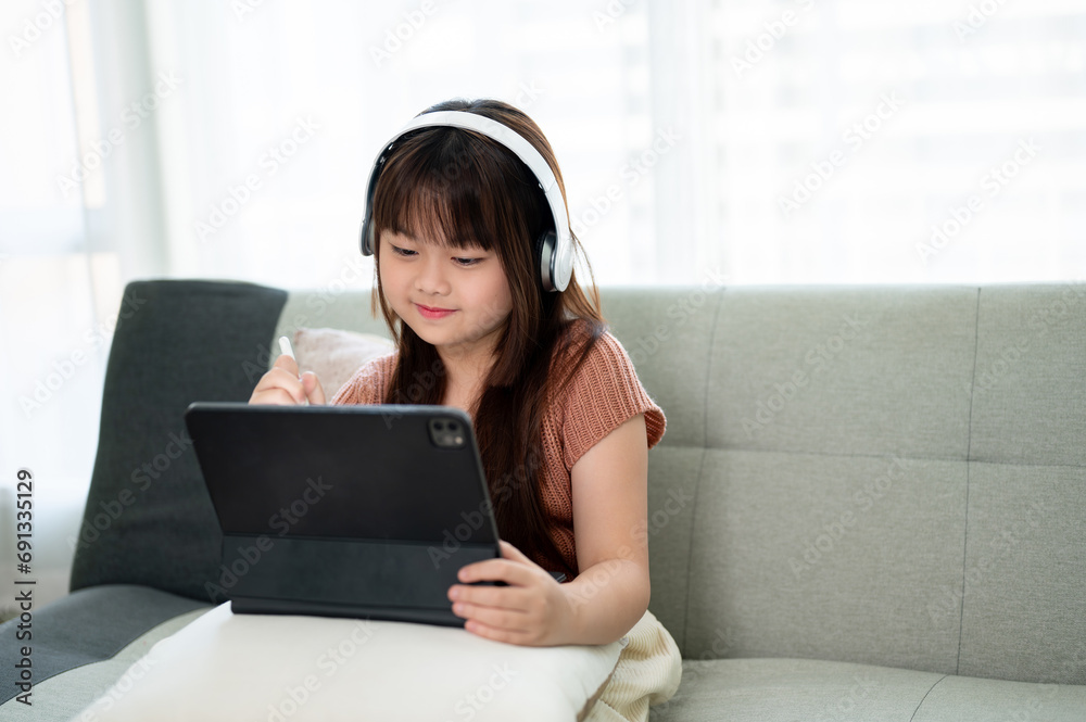 An adorable young Asian girl studying online at home on her digital tablet while sitting on a sofa. An adorable young Asian girl studying online at home on her digital tablet while sitting on a sofa.