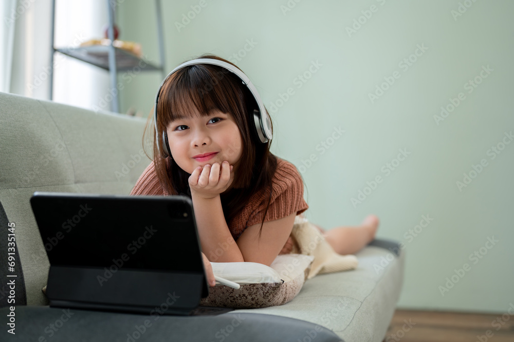 Young adorable Asian girl in headphones is lying on a sofa in the living room with her tablet. Young adorable Asian girl in headphones is lying on a sofa in the living room with her tablet.