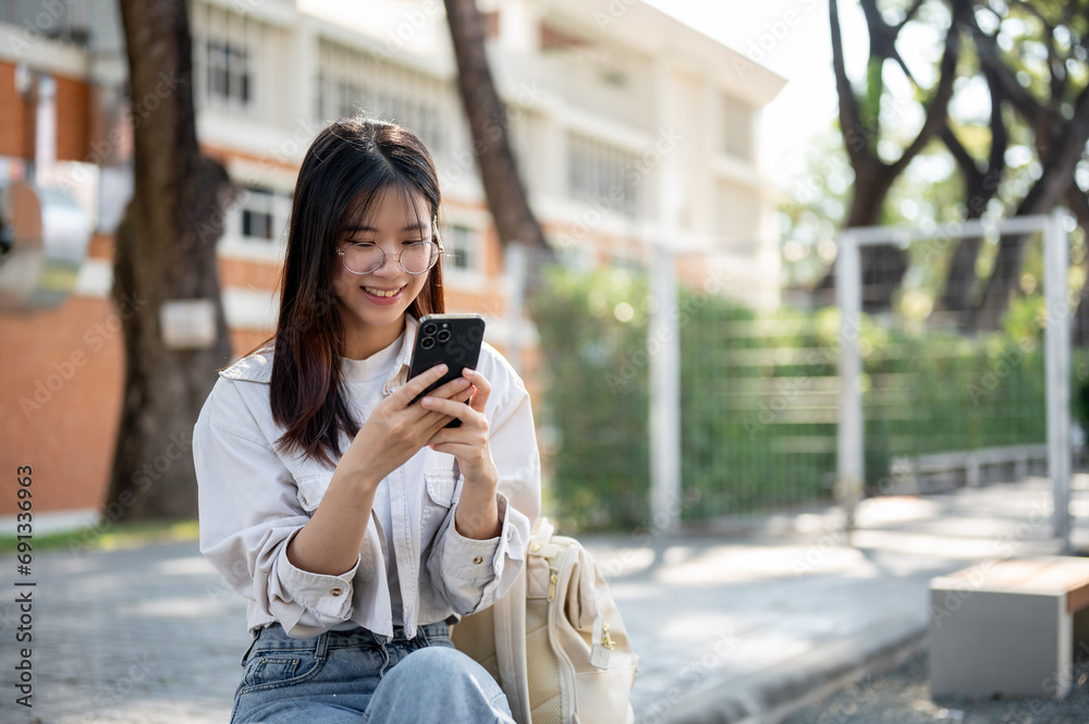 A positive Asian woman is chatting with her friends on her phone while resting on a bench in a park.