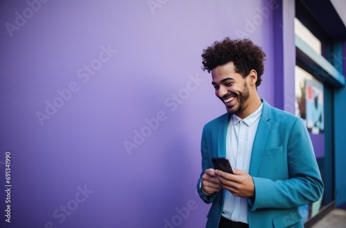 a young man is smiling and holding his smartphone next to a purple wall