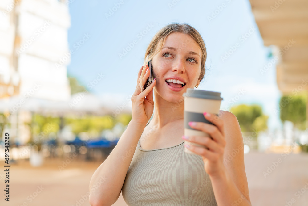 Young blonde woman at outdoors using mobile phone and holding a coffee with happy expression Young blonde woman at outdoors using mobile phone and holding a coffee with happy expression