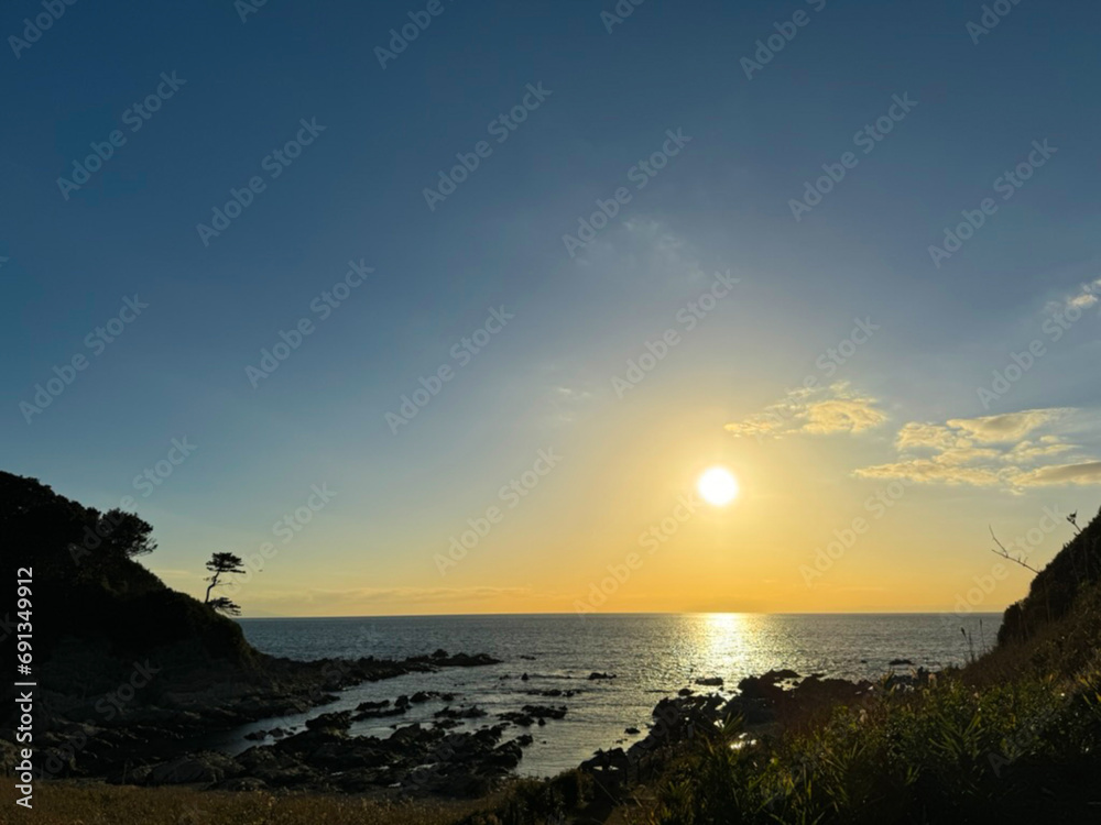 大きな海に波が打ち寄せ、青い大空と太陽と夕日が混ざり合い、高台から見下ろす実写写真