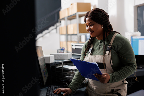 Stockroom supervisor checking clients online orders on computer before start preparing packages for shipping in storage room. African american worker wearing industrial overall standing at counter photo