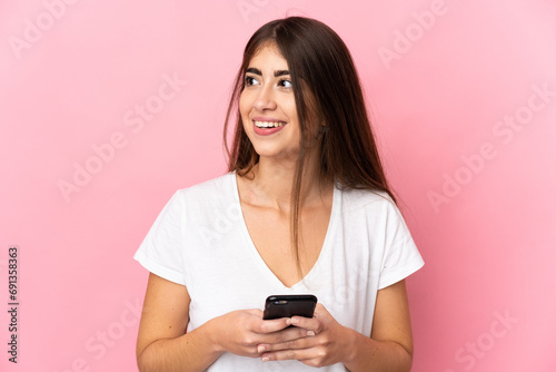 Young caucasian woman isolated on pink background using mobile phone and looking up