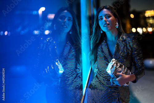 Woman holding clutch bag and standing near glass wall with neon lighting photo